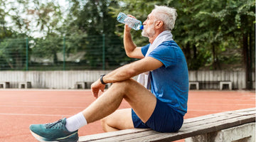 An older man in athletic clothing sits on a bench at an outdoor track, drinking from a water bottle after exercise, with trees and the track visible in the background.