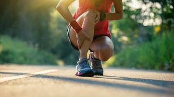 A runner kneels on a sunlit path, gripping their lower leg in apparent pain, wearing athletic shoes and workout clothing with greenery blurred in the background.