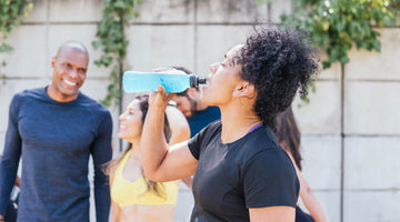 A woman in athletic clothing drinks from a blue sports bottle outdoors, while a small group of people in workout gear stand behind her, smiling and resting after exercise.