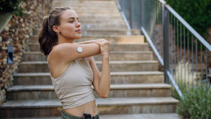 Woman stretching her shoulder outdoors on a set of stairs, wearing athletic clothing and a continuous glucose monitor on her arm, preparing for a workout.