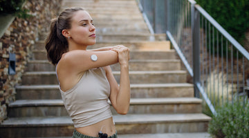 Woman stretching her shoulder outdoors on a set of stairs, wearing athletic clothing and a continuous glucose monitor on her arm, preparing for a workout.