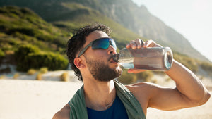 A man wearing sunglasses drinks from a water bottle outdoors, standing in a sunny, mountainous landscape, suggesting hydration during a workout or hike.
