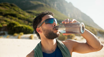 A man wearing sunglasses drinks from a water bottle outdoors, standing in a sunny, mountainous landscape, suggesting hydration during a workout or hike.