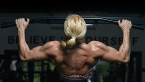 Alt text: A muscular woman seen from behind performs a pull-up on a bar in a gym, highlighting her defined back and arm muscles.