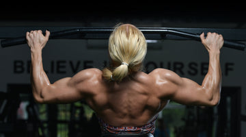 Alt text: A muscular woman seen from behind performs a pull-up on a bar in a gym, highlighting her defined back and arm muscles.