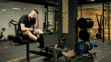 A tired man sits on a leg extension machine in a gym, resting his head in his hand, with weights and workout equipment visible around him.