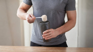 Man adding a scoop of protein powder into a shaker bottle on a table, preparing a drink for a workout or recovery.
