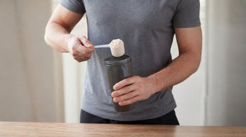 Man adding a scoop of protein powder into a shaker bottle on a table, preparing a drink for a workout or recovery.