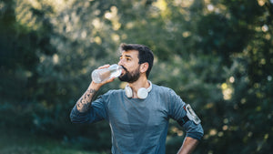 A man in athletic clothing pauses during an outdoor workout to drink from a water bottle, with headphones around his neck and trees blurred in the background.