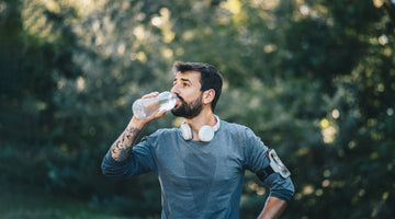 A man in athletic clothing pauses during an outdoor workout to drink from a water bottle, with headphones around his neck and trees blurred in the background.