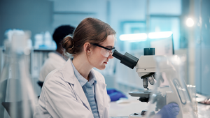 Scientist in a lab coat and safety glasses examining a sample through a microscope in a bright laboratory.