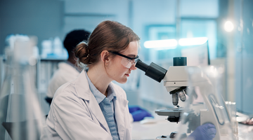 Scientist in a lab coat and safety glasses examining a sample through a microscope in a bright laboratory.